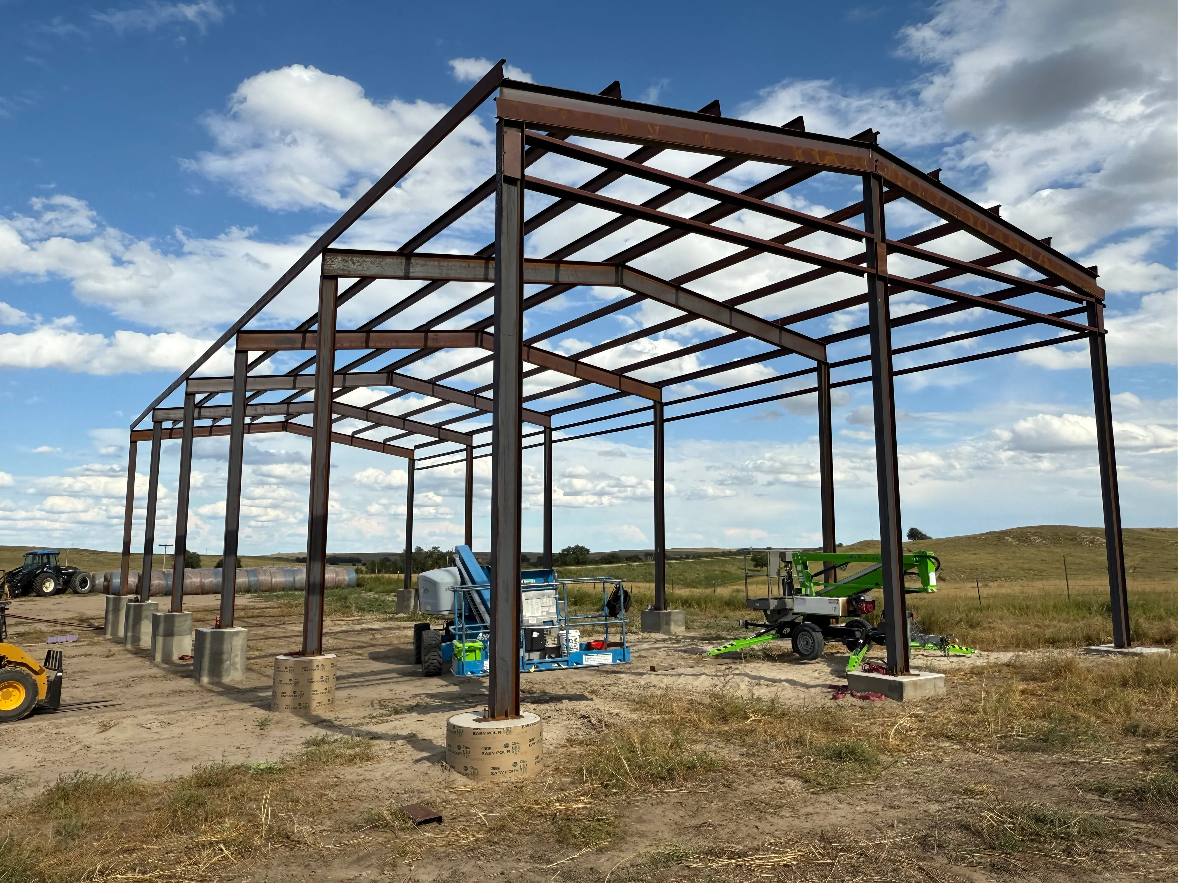 Steel I-beam frame structure with diagonal bracing and concrete footings on rural site