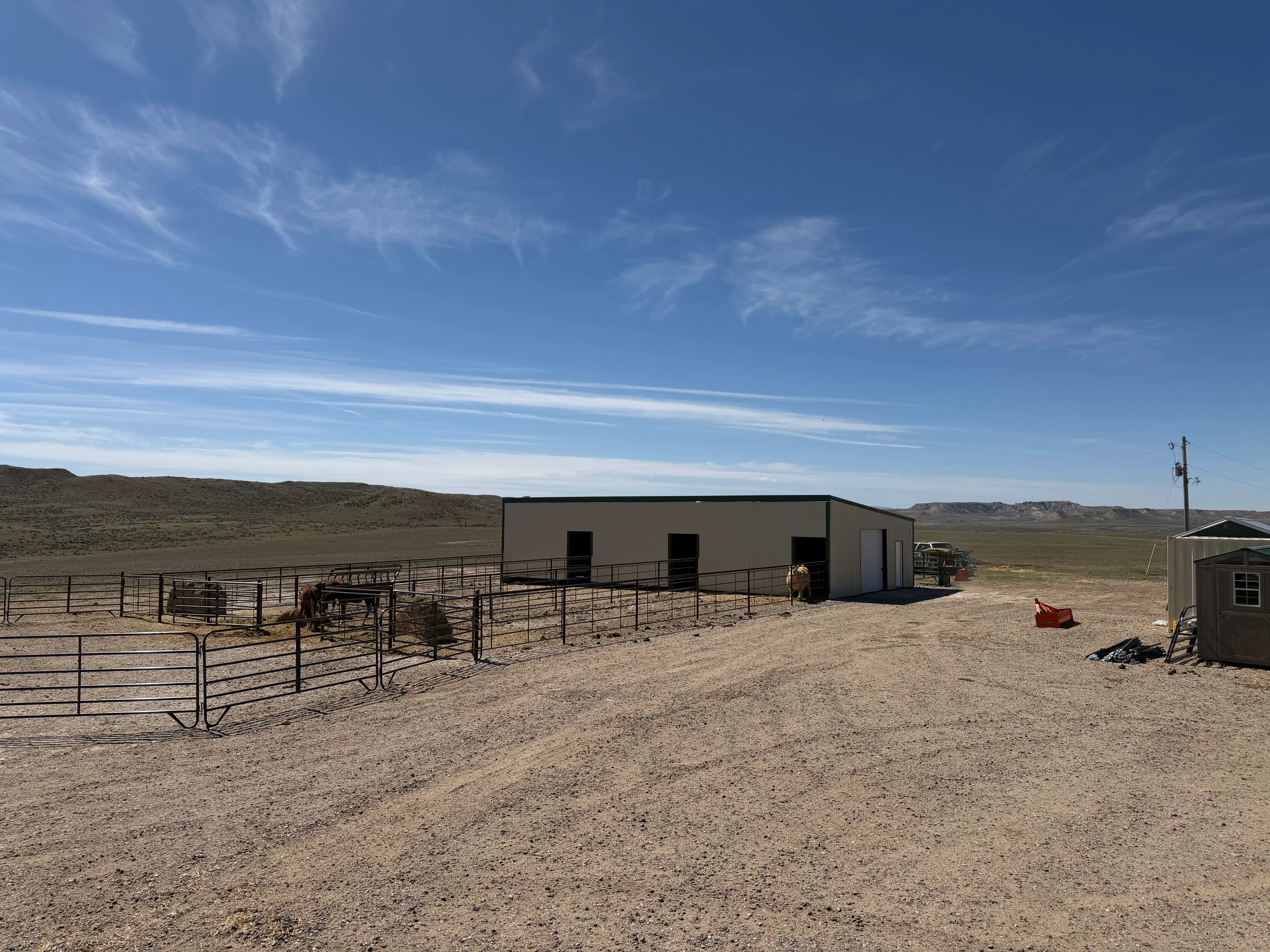 Tan metal agricultural building with steel pipe corrals and livestock gates on dirt lot