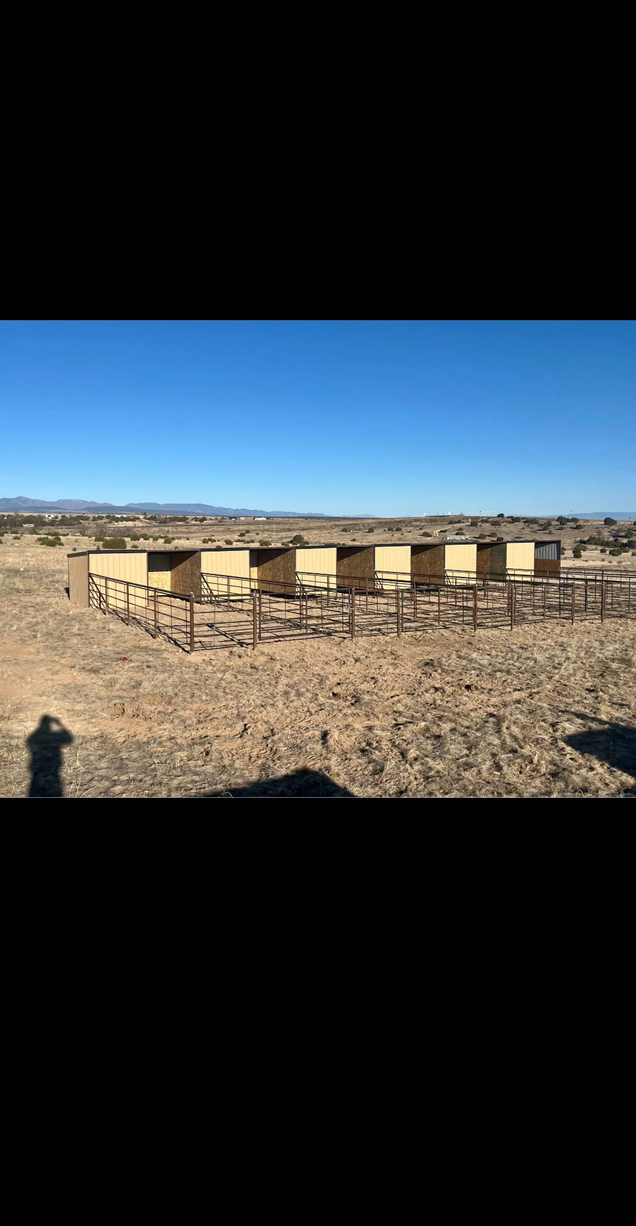 Eight tan corrugated metal livestock shelters with steel pipe fencing and gates on dirt ground
