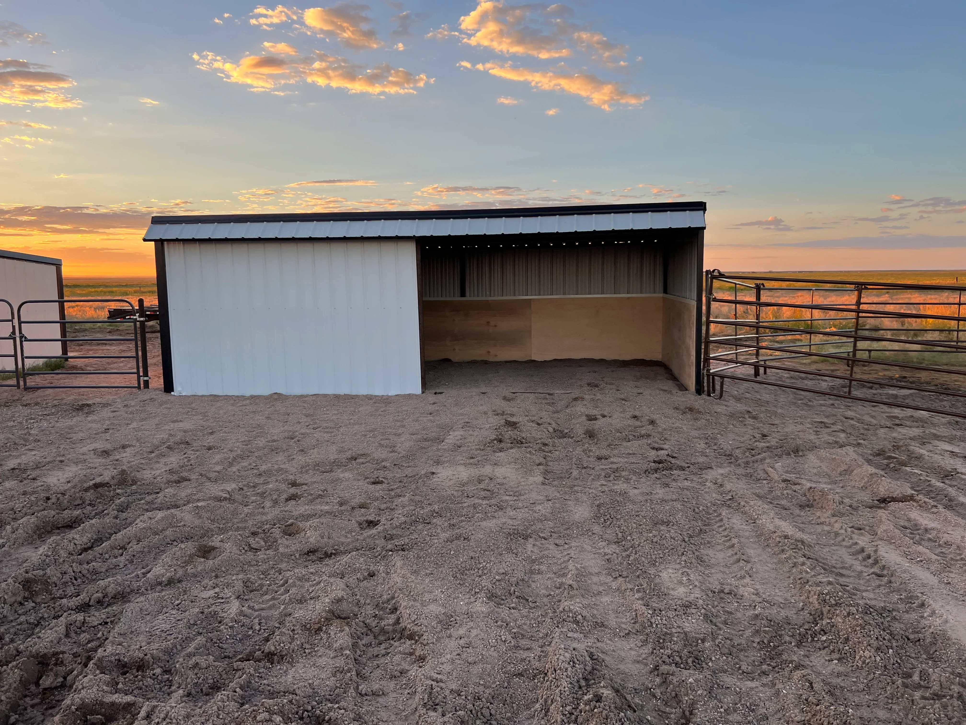 White corrugated metal building with open lean-to shelter, steel gates, concrete pad, and fencing
