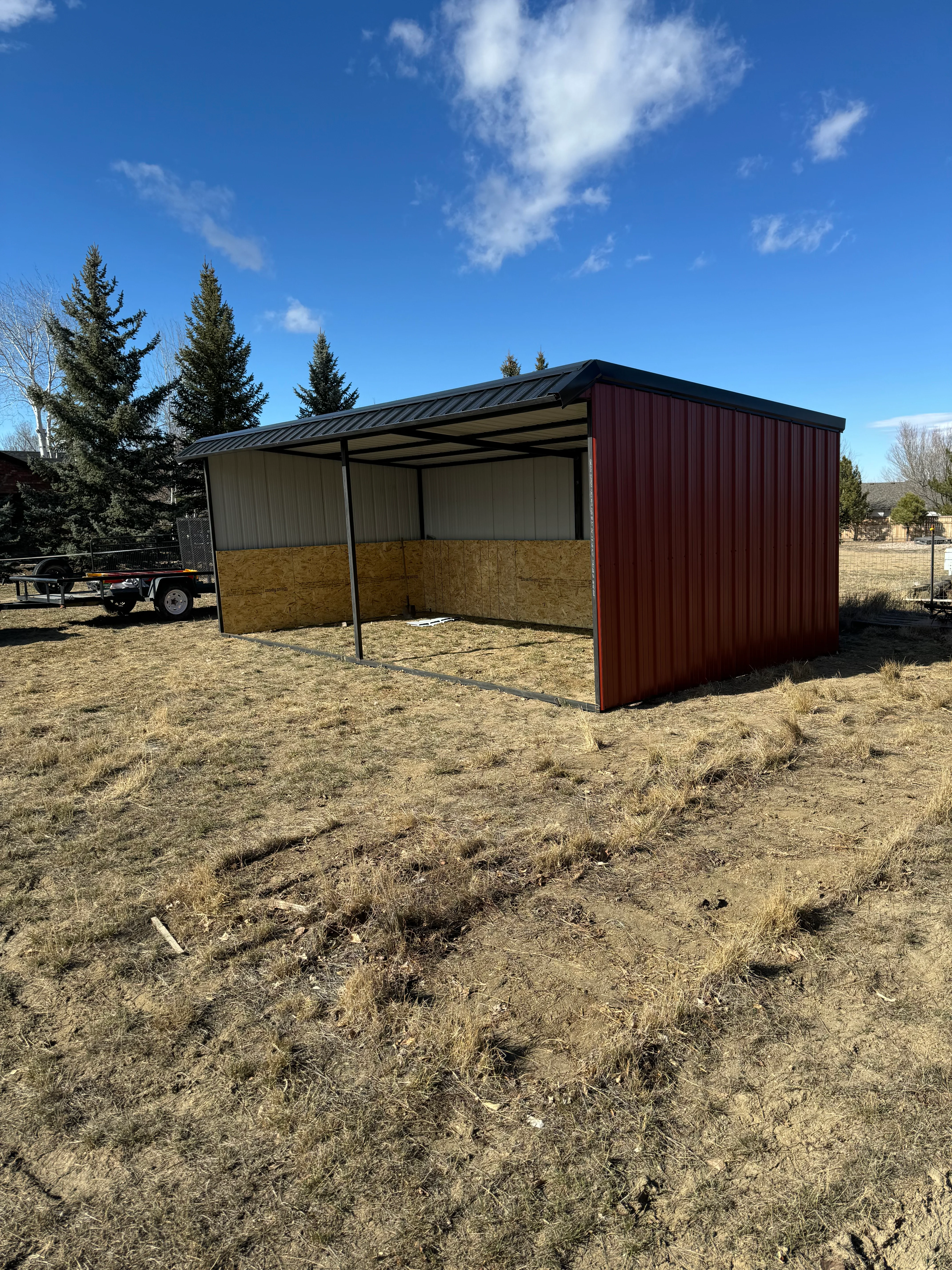 Metal loafing shelter with open sides and attached burgundy storage box in rural setting