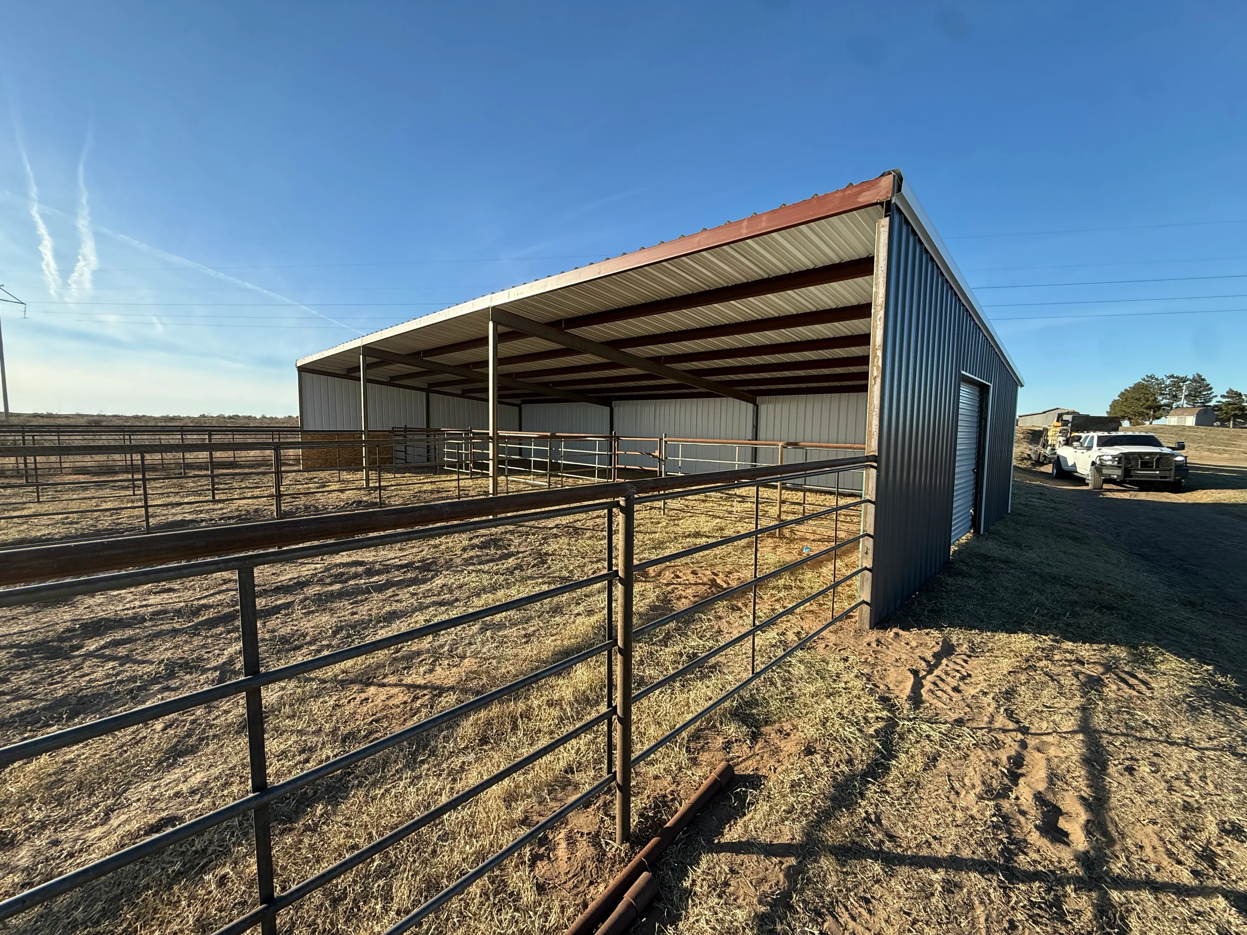 Metal frame shelter with corrugated roof, steel pipe fencing, and enclosed utility building on rural property