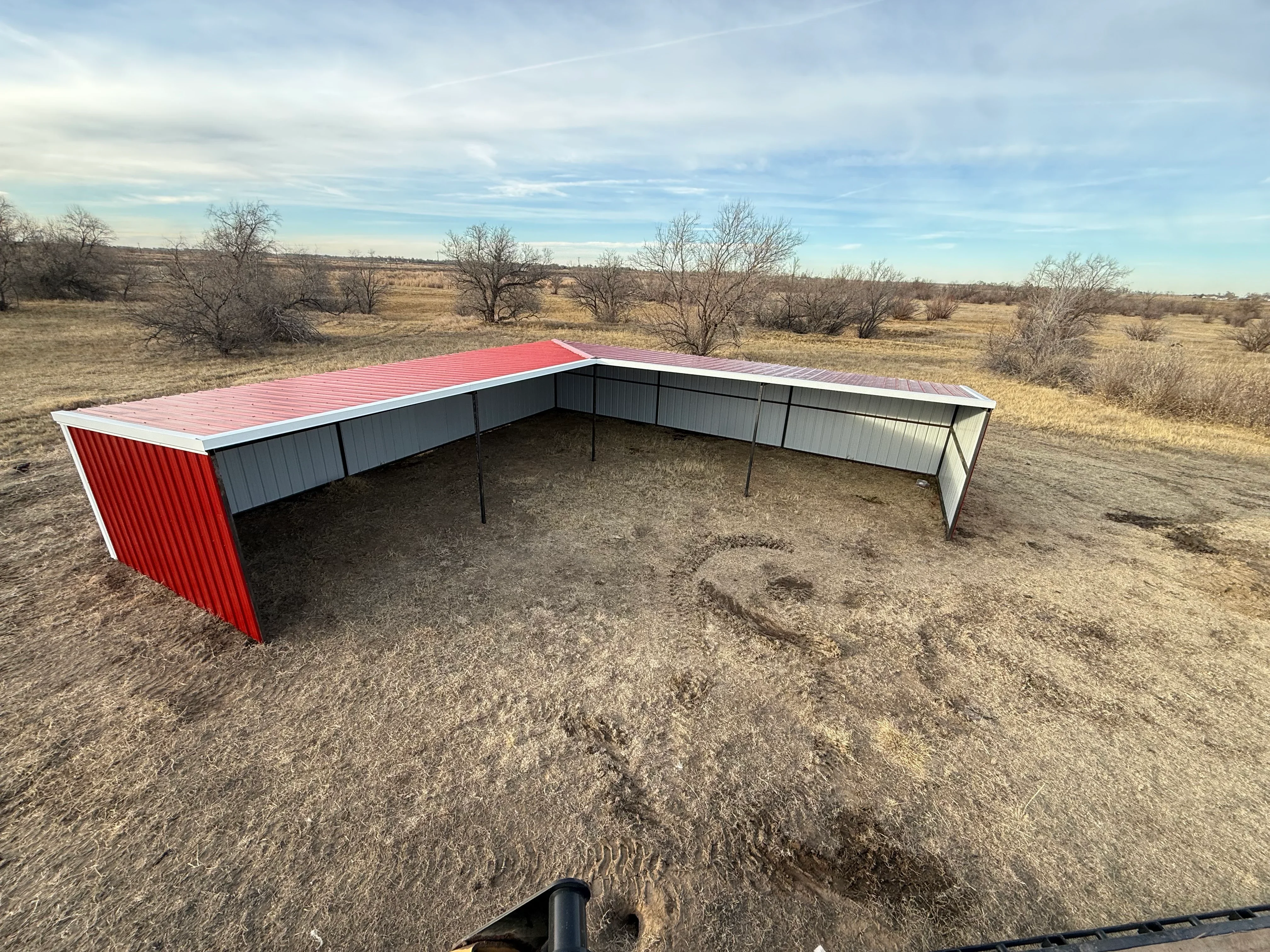 L-shaped metal shelter with red and gray corrugated steel panels and open sides on dirt ground