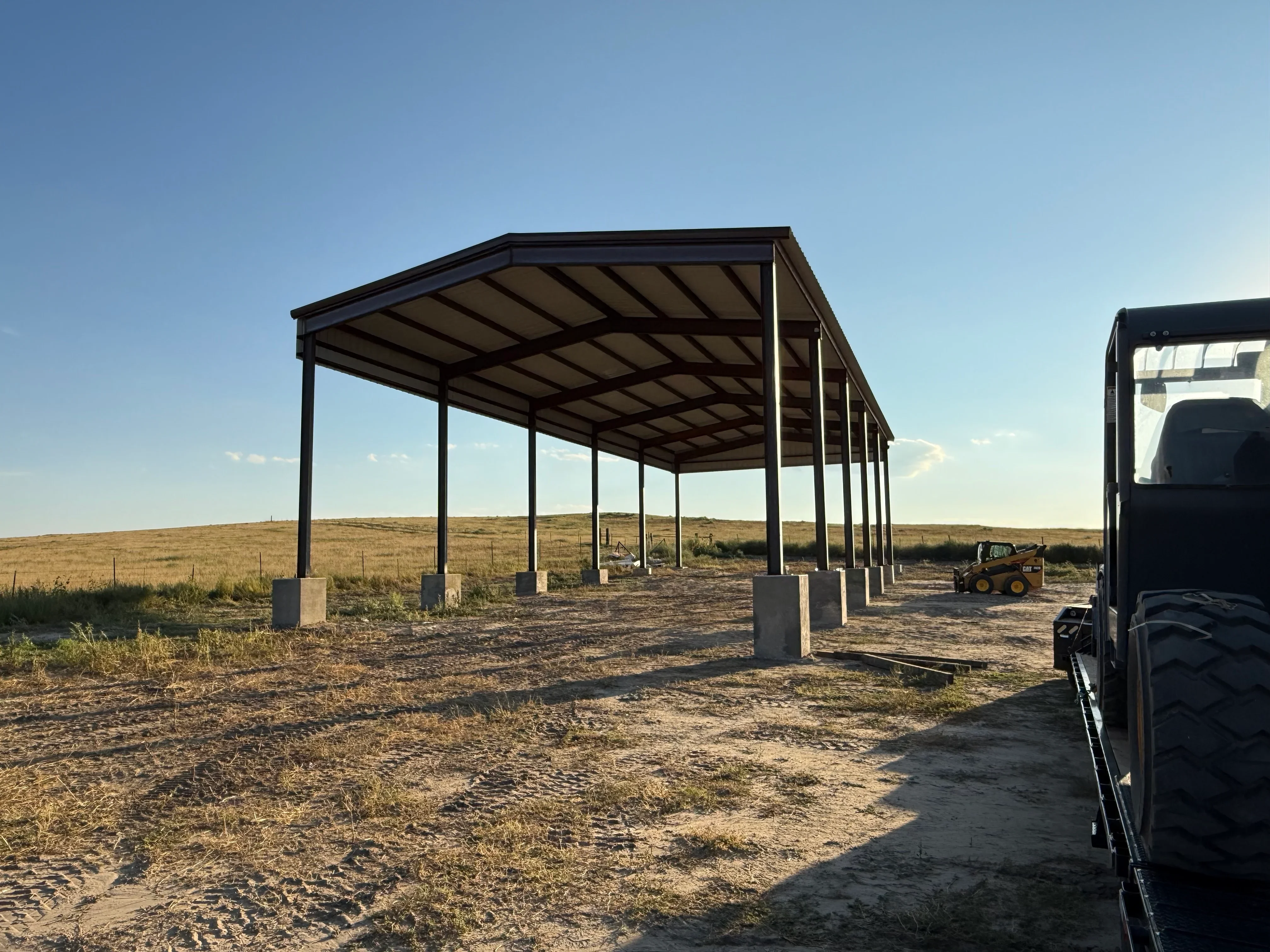 Steel frame carport with black columns, angled metal roof panels, and concrete footings on rural property