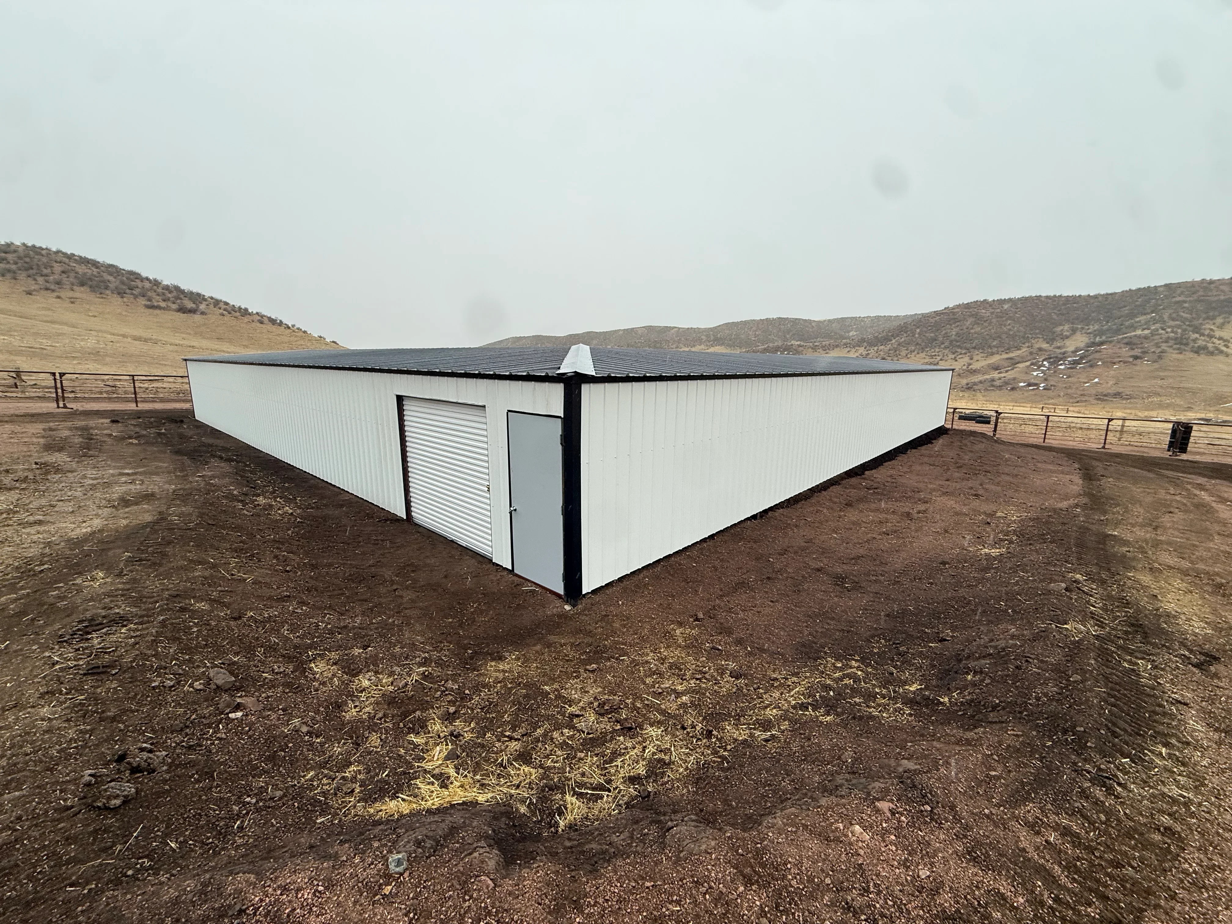 White corrugated metal building with black frame, louvered vent, and entry door on dirt foundation