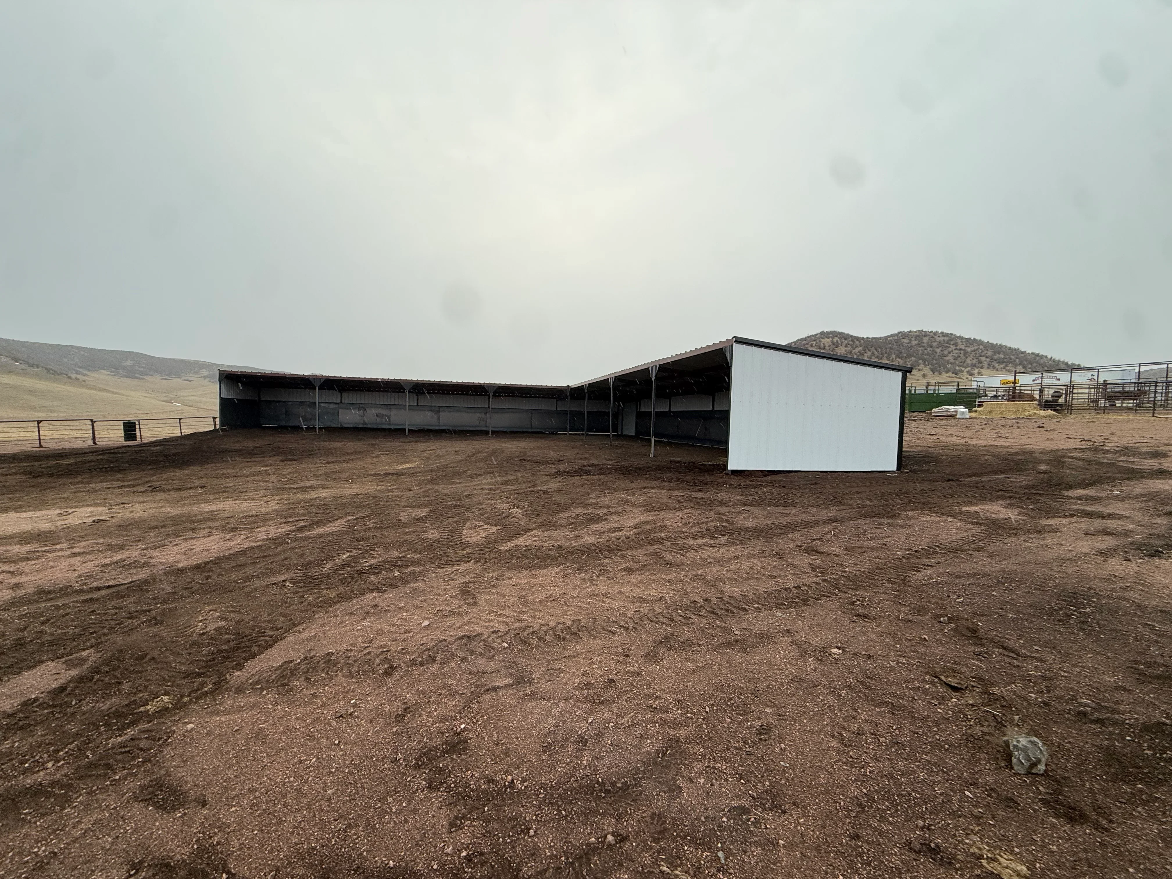 Black steel-framed agricultural shelter and white metal building on prepared site with fencing
